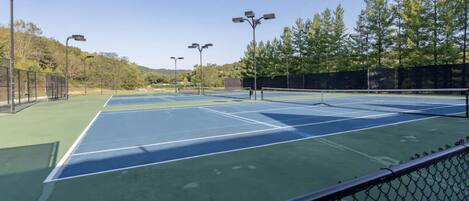 Canchas de tenis múltiples, rodeadas por una cerca de malla metálica y equipadas con iluminación aérea.