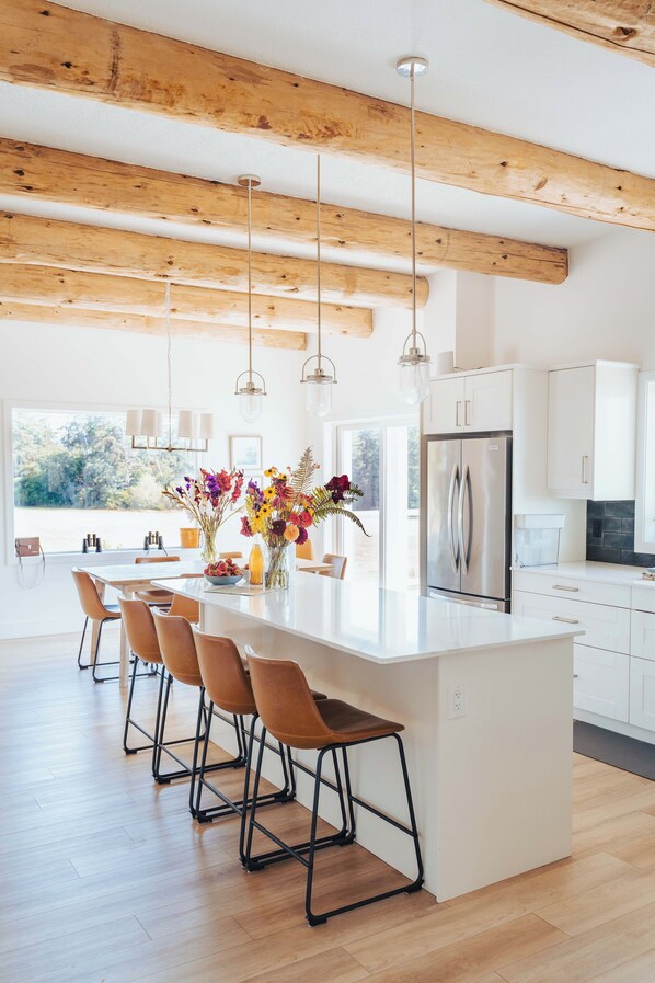 Cocina moderna con una isla grande, vigas de madera, luces colgantes y vistas al exterior.
