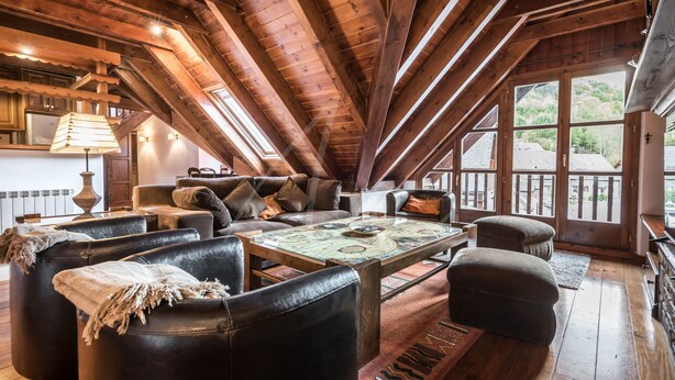 A cozy loft-style room with a high ceiling, wooden beams, a large window, and a seating area with leather sofas and ottomans. - Valartíes (Artíes)