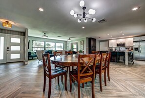 Un grand espace à manger avec une table et des chaises en bois, une cuisine équipée d’appareils électroménagers modernes et un salon avec un canapé et un ventilateur de plafond.