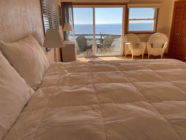 A bedroom with a large bed, a view of the sea through a sliding glass door, and a balcony with chairs.