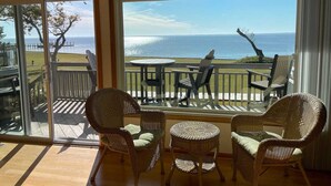 Quarto com cadeiras de vime e uma pequena mesa, janelas amplas com vista para o oceano e área de deck com cadeiras e mesa.