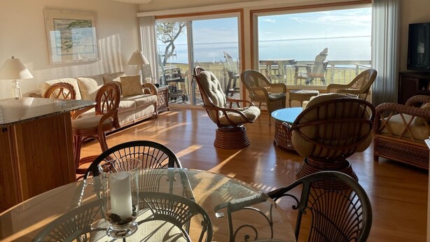 Uma sala de estar ampla, com uma grande janela com vista para o oceano, mobiliada com cadeiras de vime e uma mesa de jantar de vidro.