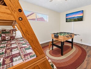 A bedroom with a bed, a wooden headboard, a window with blinds, a wall-mounted TV, and a foosball table. - Knotty Hideawa - Foosball, Fireplace, Deck, Fire Pit, BBQ (Big Bear City)