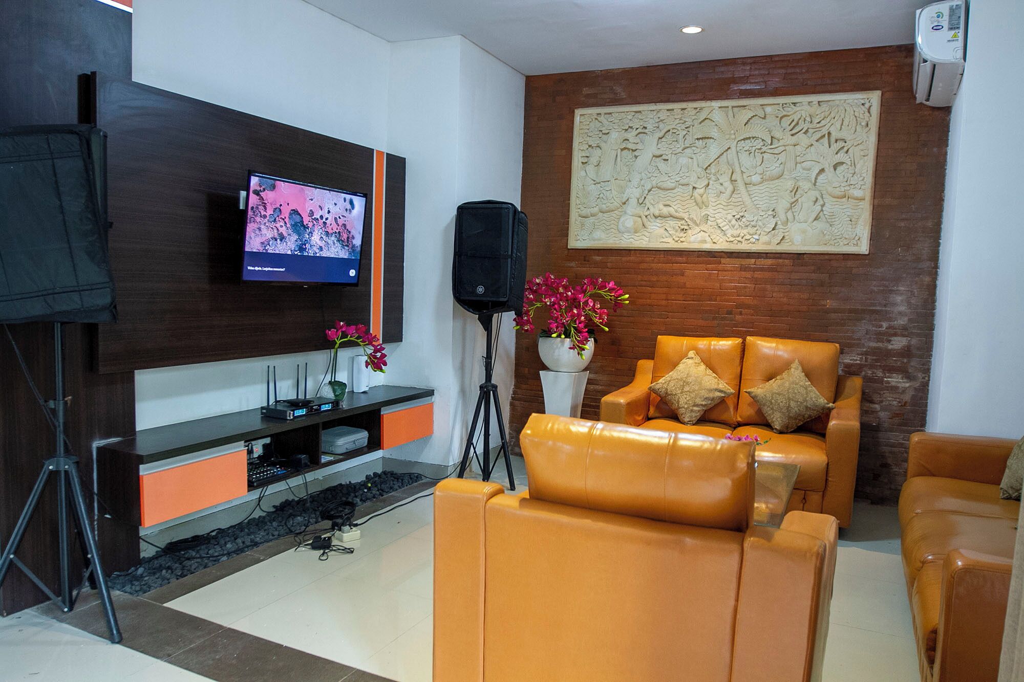 A modern living room with a leather sofa, a flat-screen TV mounted on a dark wood panel, a large decorative wall piece, and a potted plant with pink flowers.