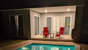 Ein Pool mit zwei roten Stühlen und einem Tisch, umgeben von einer überdachten Terrasse mit Fenstern und Vorhängen.