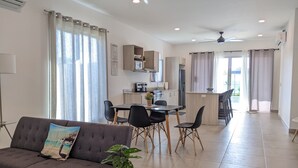 Ein modernes Wohnzimmer mit einer Couch, einem Esstisch und Stühlen. Im Hintergrund ist eine Küchenzeile mit Schränken und einem Kühlschrank zu sehen.