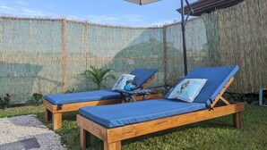 Ein Garten mit zwei Holzliegen unter einem großen Sonnenschirm, einem Bambuszaun und einer kleinen Zimmerpalme.