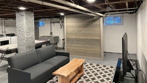 A basement with exposed pipes, a gray sofa, a wooden coffee table, a television, and a patterned rug. - New Spacious single w/ parking.  Bikes, XBOX, (Phoenixville)
