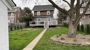 A residential backyard with a paved pathway leading to a deck, surrounded by a brick house and a tree.