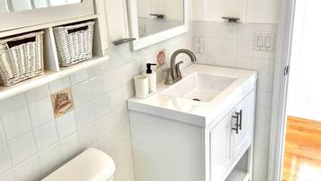 Ein Badezimmer mit Toilette, Waschbecken und Spiegel. Wickerkörbe befinden sich auf einem Regal und ein kleines dekoratives Element ist an der Wand angebracht.