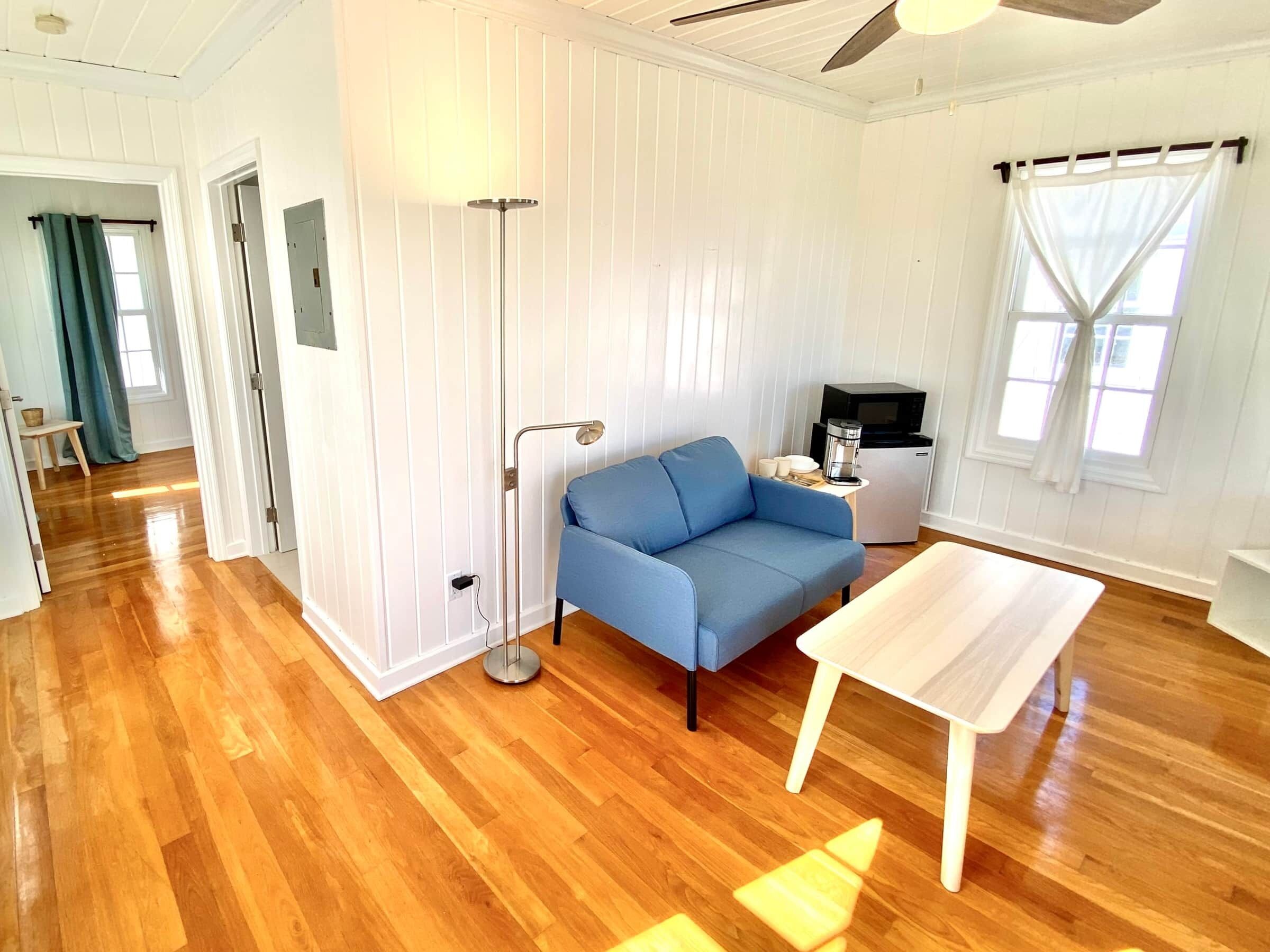 Une pièce avec un canapé bleu, une table basse blanche, un lampadaire, une petite table sur laquelle se trouve une machine à café et un four à micro-ondes.