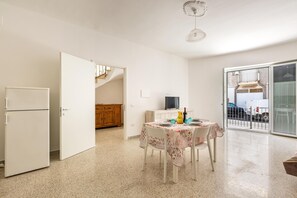 A dining area with a table set for two, a refrigerator, a TV, and a sliding glass door leading to a balcony.