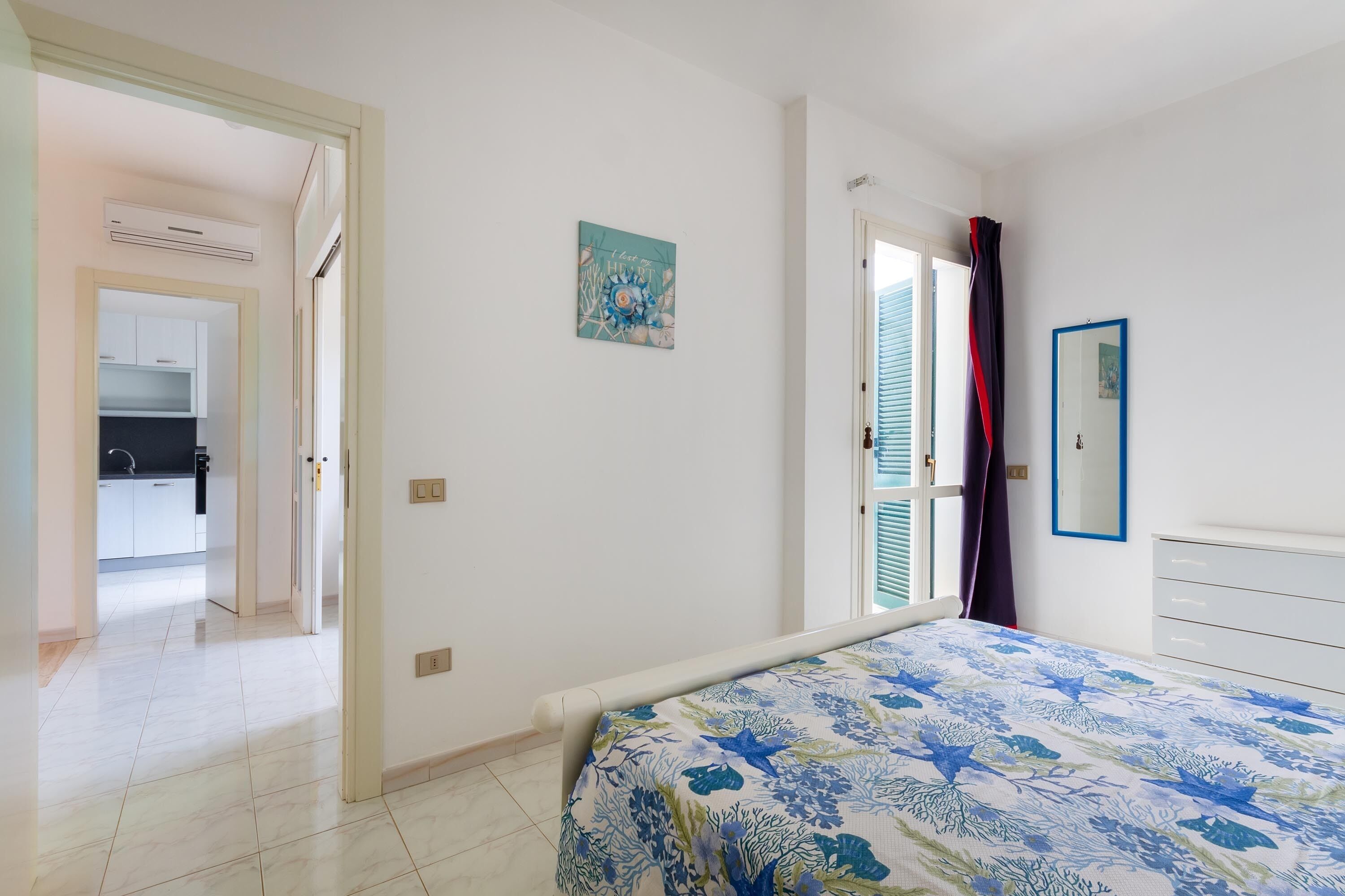 Une chambre à coucher avec un lit, une porte donnant sur une cuisine et une fenêtre avec des rideaux.