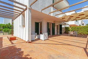 Une vaste terrasse extérieure dotée d’un sol carrelé, d’un espace salon avec des meubles blancs et d’une partie couverte avec des poutres en bois.