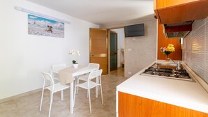 A small kitchen with a gas stove, a white table with two chairs, and a painting on the wall. - 2904 Residence Mareluna - Bilo 2 by Barbarhouse (Porto Cesareo)