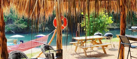 An outdoor seating area with wooden tables and chairs, a hammock, and a view of a body of water.