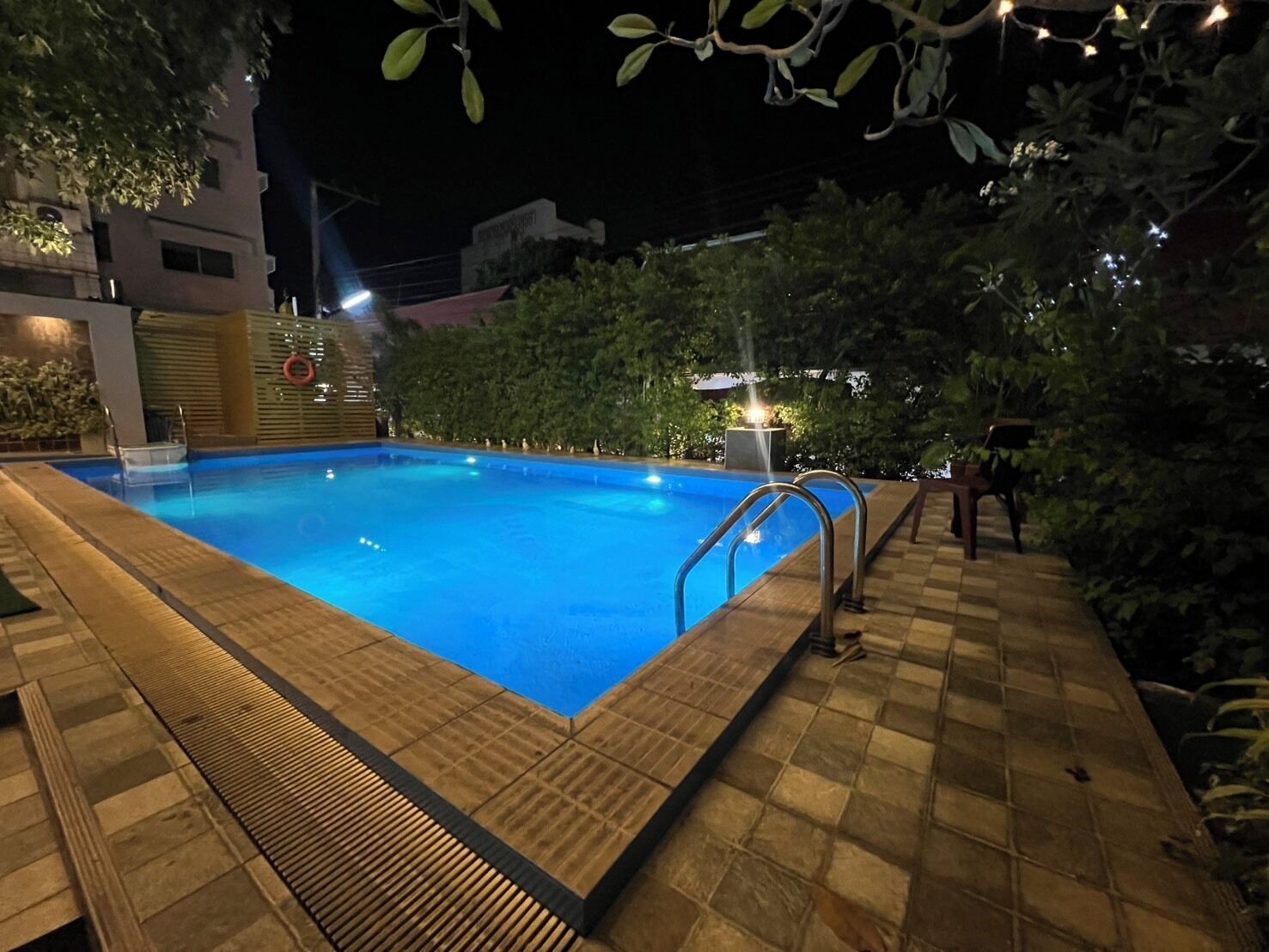 Una piscina con plataforma de azulejos, rodeada de vegetación y un edificio al fondo.