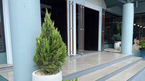 Entrada de prédio com uma árvore sempre-verde em vaso, um vaso de samambaia e uma porta de vidro que dá para o interior.