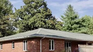 Een laag huis met bakstenen muren, een schuin dak, een voortuin met gazon en enkele potplanten, en een veranda.
