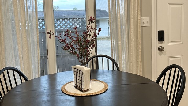 Een eetgedeelte met een donker houten tafel, vier stoelen, een decoratieve vaas met rode bloemen en een bordje erboven met de tekst 'GATHER 'ROUND the table'.