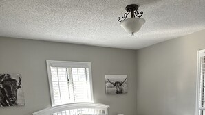 Een slaapkamer met een wit bed, twee nachtkastjes, een ventilator in het plafond en een raam met jaloezieën.