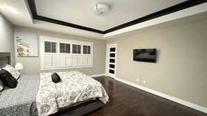A bedroom with a bed, a TV, a window with shutters, and a ceiling light.