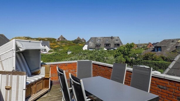 A rooftop patio with a table and chairs, a grill, and a view of houses with thatched roofs.
