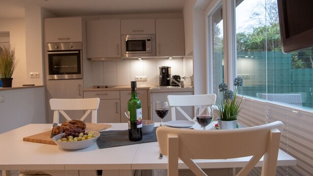 Una cocina moderna con armarios blancos, una mesa de comedor con sillas, una botella de vino y un bol de fruta.
