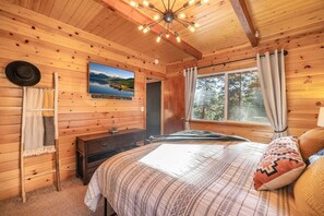 A cozy cabin bedroom with a wooden interior, a bed with patterned bedding, a television on the wall, and a window with curtains. - MODERN RUSTIC CABIN W/SPA, WALKING DISTANCE TO SKI RESORT, ZOO & GOLF COURSE (Big Bear Lake)