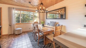 Un coin repas accueillant avec une table en bois, des chaises et un lustre. On y trouve une grande fenêtre avec des rideaux, un miroir au mur et un petit tabouret.