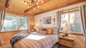 Une chambre confortable, aménagée dans un chalet en bois, avec un lit, des tables de chevet et une grande fenêtre agrémentée de rideaux.