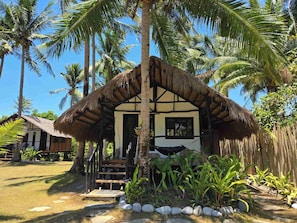 A thatched-roof hut with a wooden staircase leading to a door, surrounded by palm trees and a garden with rocks and plants. - Whole Resort (Binga)
