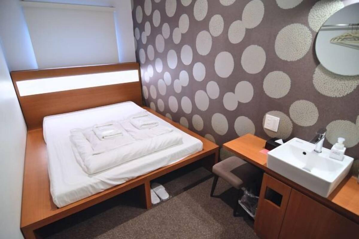 A single bed with a wooden headboard, a bedside table, a small stool, a bathroom with a sink and mirror, and a polka dot wall.
