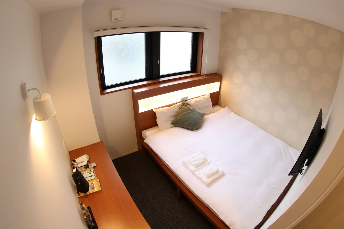 A small, neatly made bed with white linens and two pillows, a bedside table with a lamp and a remote control, and a window with blinds.