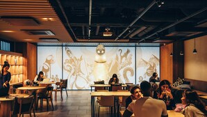 Un interior moderno con mesas y sillas de madera, una pantalla de proyección con diseños abstractos y un techo con conductos expuestos.