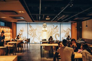 A modern interior with wooden tables and chairs, a projection screen with abstract designs, and a ceiling with exposed ductwork. - WeBase KYOTO - Hostel (Kyoto)