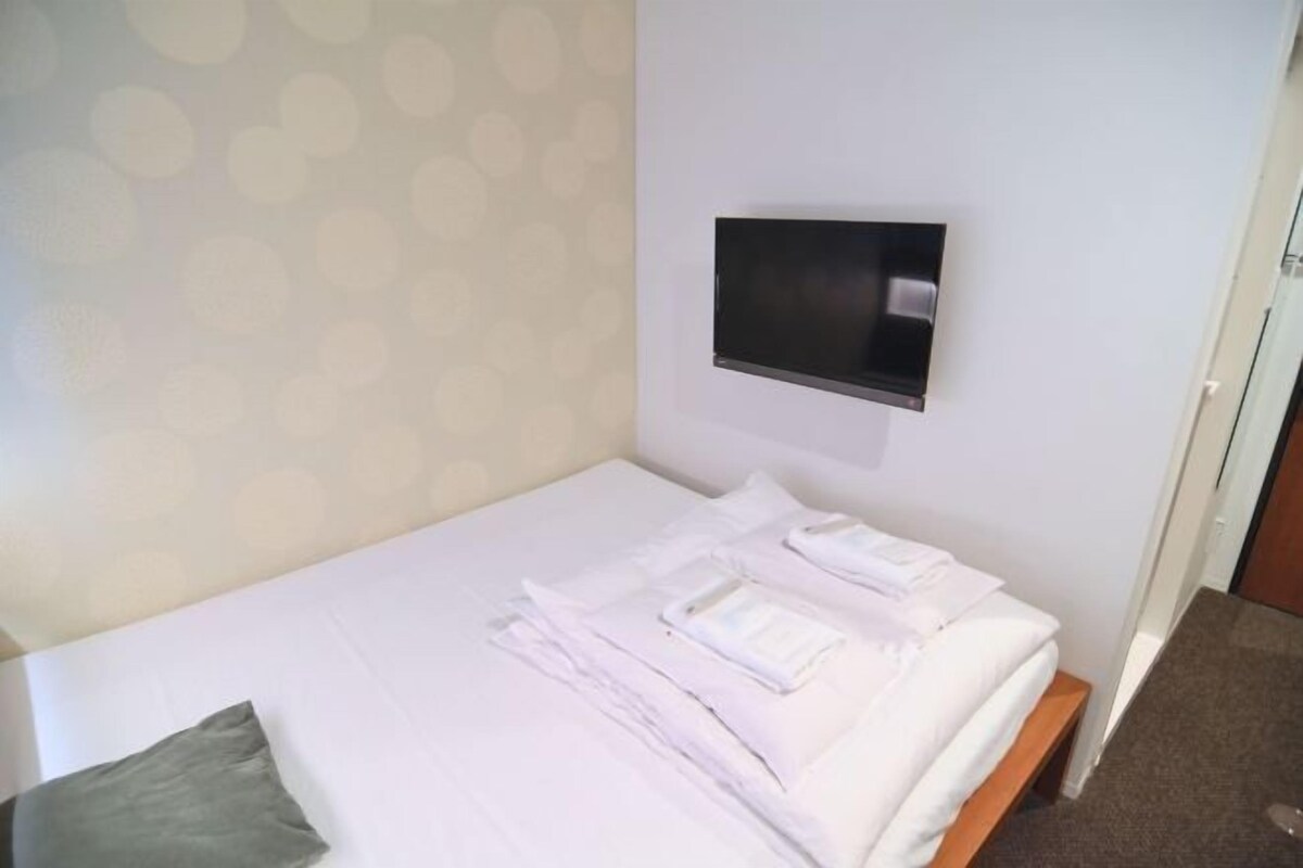 A single bed with neatly folded white linens, a mounted TV on the wall, and a patterned wall.
