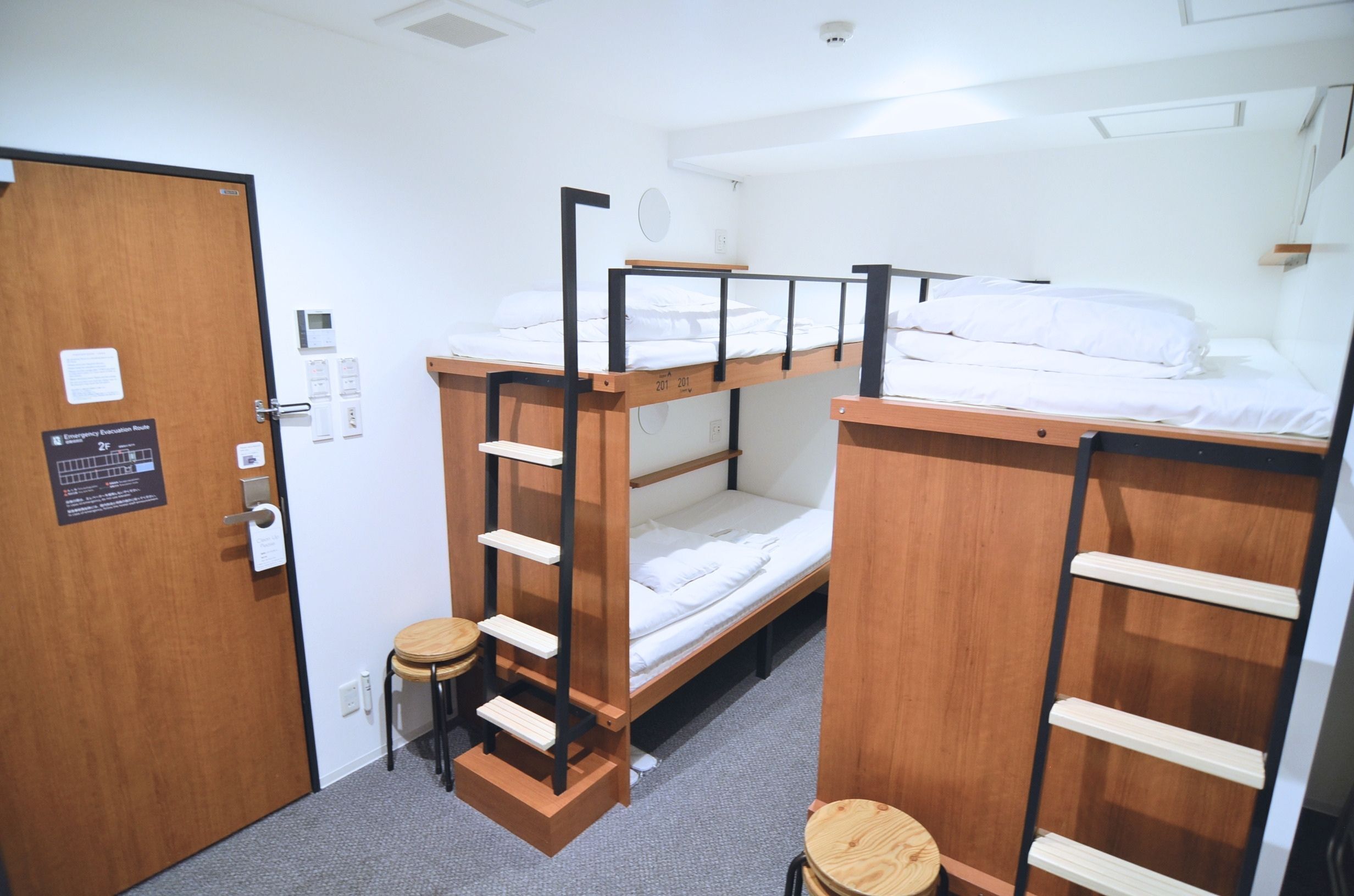 Habitación con literas, una puerta, un taburete pequeño y una estantería fijada a la pared.