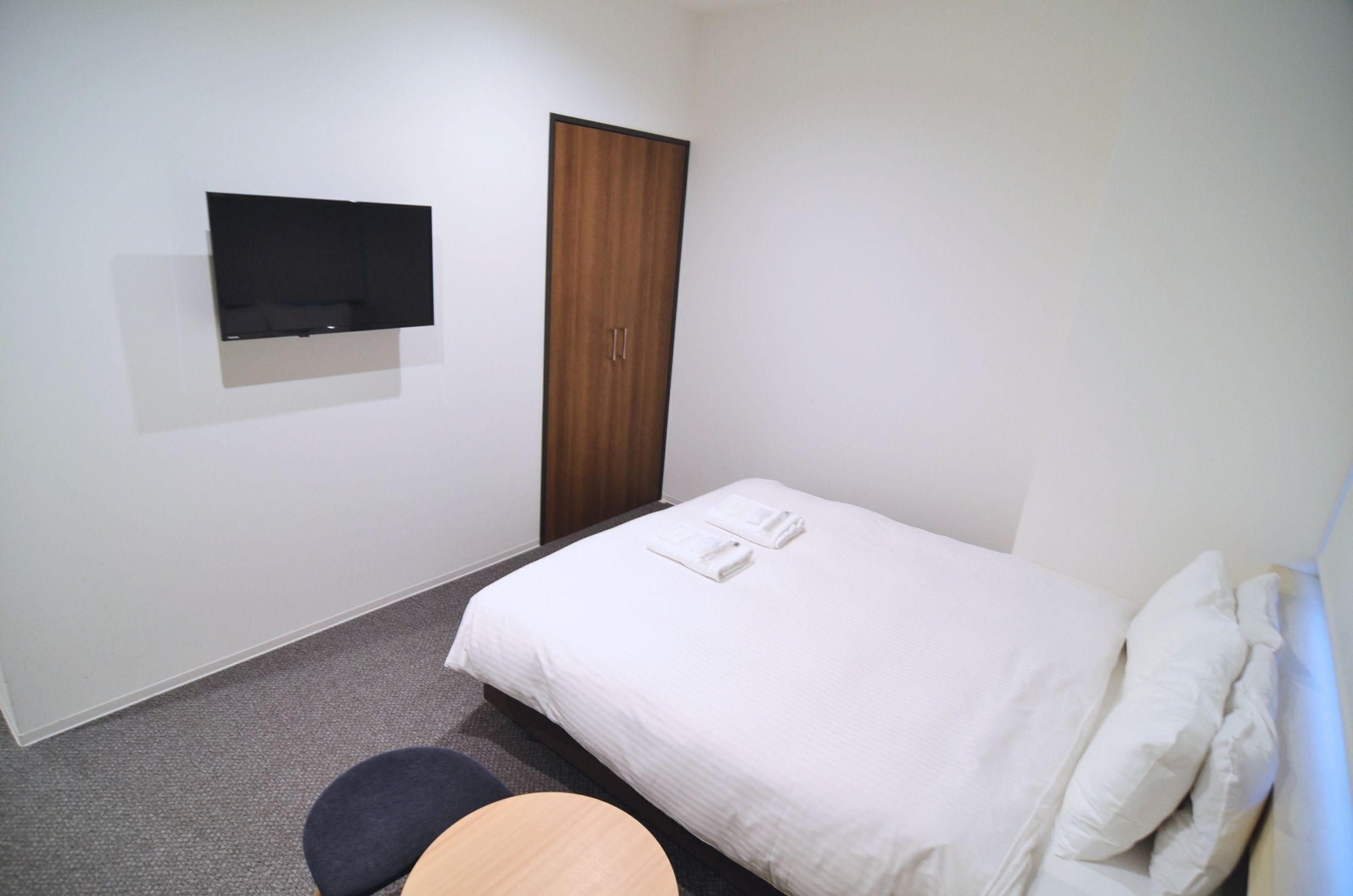 Habitación de hotel con cama, televisor montado en la pared, armario y una pequeña mesa redonda.