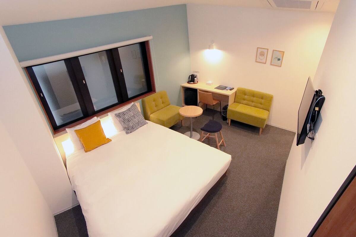 A compact hotel room with a bed, a desk, a yellow armchair, and a small round table.