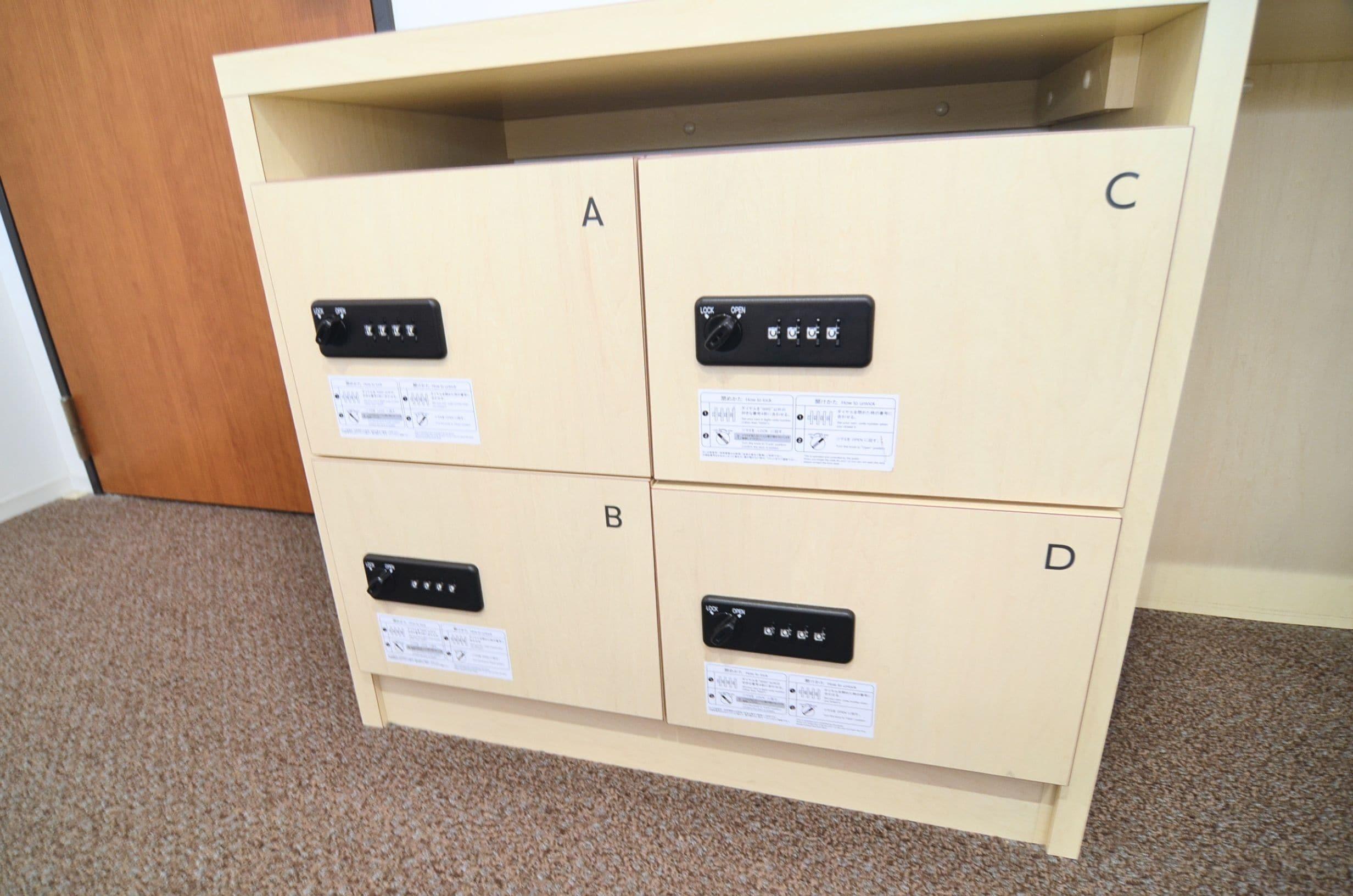 Un mueble de madera con cuatro compartimentos etiquetados A, B, C y D, cada uno con un pomo negro y una etiqueta de advertencia.