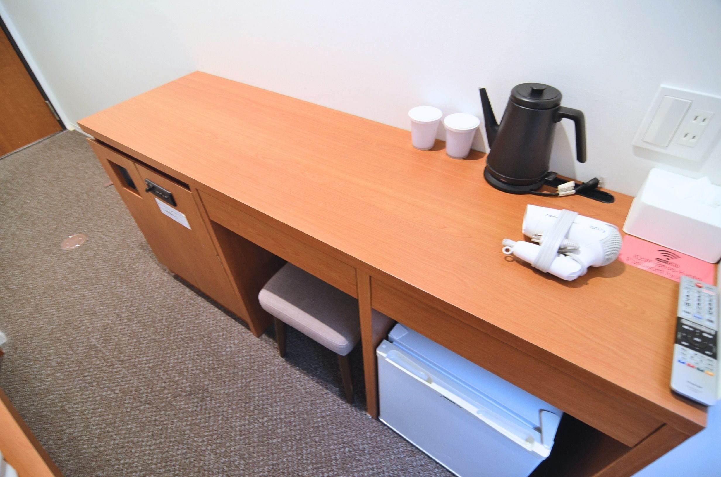 A wooden desk with a built-in mini-fridge, a kettle, and a remote control.