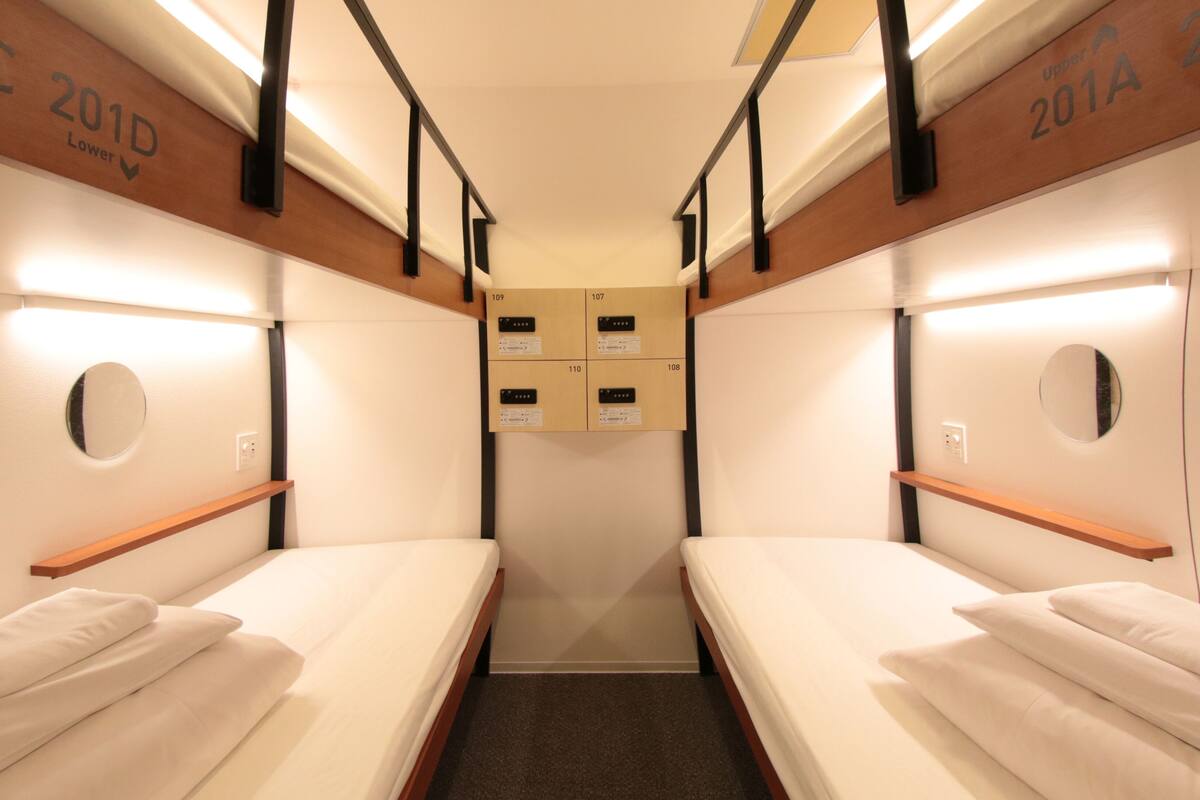 A narrow, well-lit corridor with two single beds on either side, each with a wooden shelf and a circular mirror above.