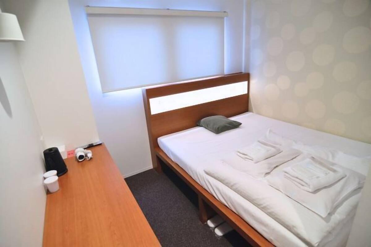 A single bed with a wooden headboard, a bedside table with a lamp, a window with blinds, and a patterned wall.