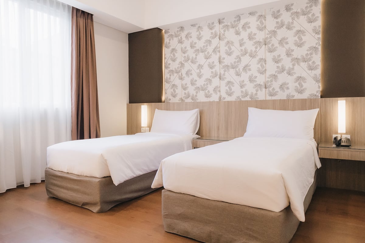 A hotel room with two beds, a wooden headboard, a floral-patterned wall, and a bedside table with a lamp.