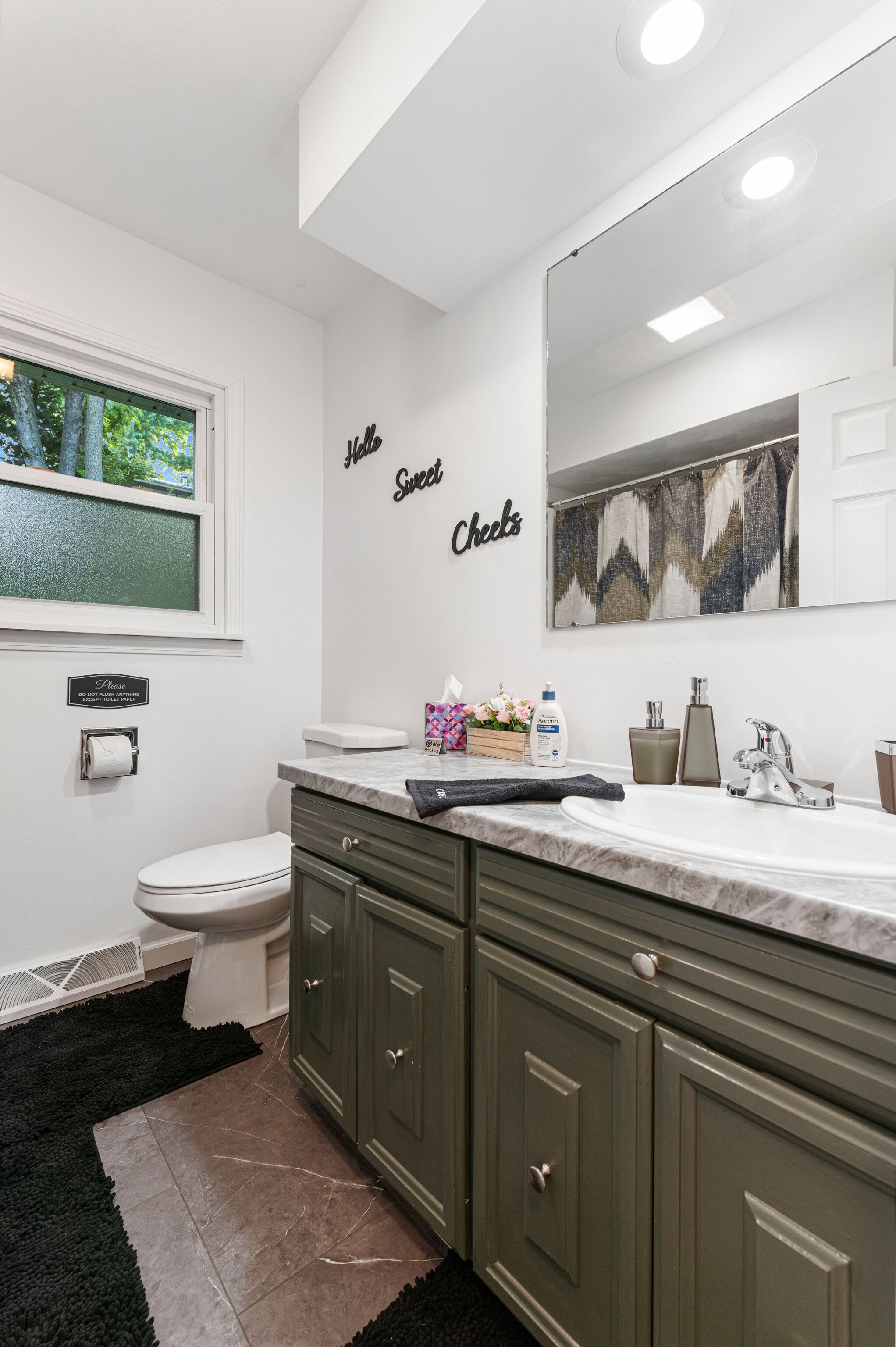 Un baño moderno con inodoro blanco, armarios verdes, un espejo grande y una ventana con vistas a árboles.