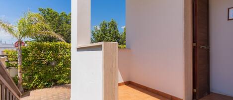 Un balcon doté d’un sol carrelé, d’une porte en bois et offrant une vue sur la verdure et un ciel dégagé.