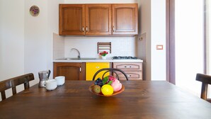 A dining table with a fruit bowl, wooden chairs, a coffee maker, and a yellow dishwasher in a kitchen with wooden cabinets and a white countertop.