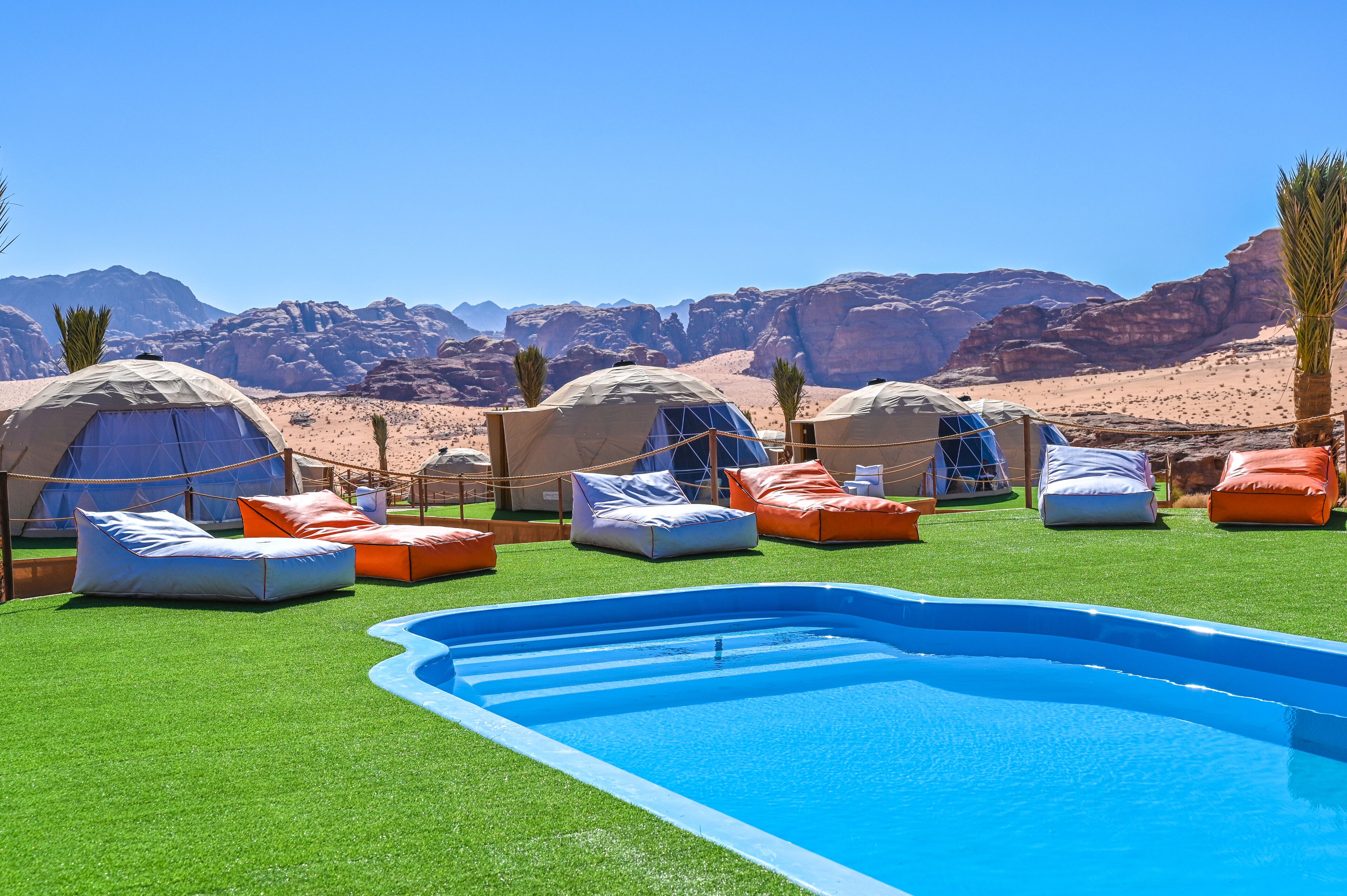 A pool area with artificial grass, several yurts, and orange cushions.